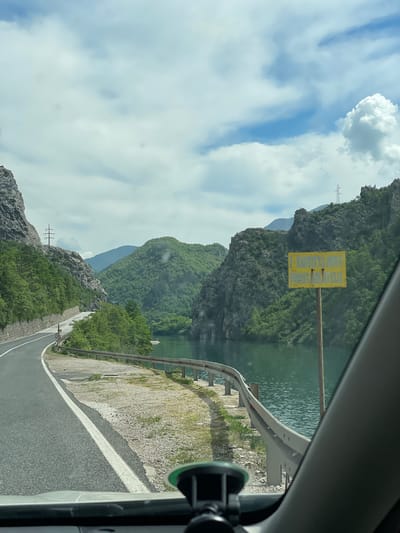 The image captures a view from inside a vehicle, looking out onto a winding road in Glogošnica, Bosnia and Herzegovina. The road is bordered by a safety guardrail on the right and is adjacent to a body of water, likely a lake or river. Lush greenery and rocky cliffs characterize the surrounding landscape. The sky is filled with a mix of blue patches and fluffy white clouds. A yellow sign is visible on the left side of the road, affixed to a post. A suction-cup-mounted device is attached to the inside of the vehicle window. The scene suggests a scenic drive through a mountainous and forested region.