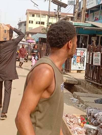 A street scene unfolds with several individuals visible. A man in a brown suit carries a box on his head, walking to the left. Another man, wearing a maroon shirt and shorts, walks in the same direction. A woman in a black shirt and patterned pants is also present. In the foreground, a man wearing a green tank top glances to the right. A street vendor's stall with various packaged goods and bottles is in front of him. The setting appears to be a busy urban area with buildings, utility poles, and open drainage.