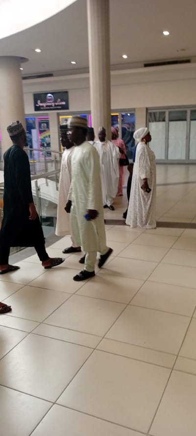The image shows a group of people walking in a shopping mall in Gandu Albasa, Nigeria. The mall has a modern design with tiled floors and large pillars. The people are dressed in traditional Nigerian attire, with men wearing kaftans and caps, and women wearing headscarves and long dresses. The people appear to be walking towards the right side of the image. There are shops in the background, with signs and displays visible. The lighting in the mall is bright and even.