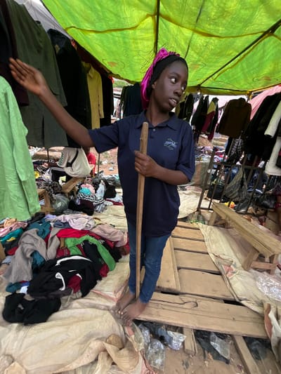 A young person, likely a vendor, lies on their back under a bright green tarp, holding a wooden pole. They are wearing a dark blue polo shirt with a visible logo and blue jeans. Their head is tilted up, looking towards the sky. The scene appears to be a market stall in Nyanya, Nigeria. Piles of colorful clothing are scattered around, some neatly stacked and others in disarray. Wooden planks and makeshift shelving are visible, suggesting a rustic and perhaps temporary setup. The overall impression is one of a busy, possibly dusty, open-air market. The lighting suggests it could be daytime, and the presence of outdoor elements hints at fair weather. The young person's expression is difficult to discern, but their posture suggests a moment of rest or perhaps engagement with their merchandise.