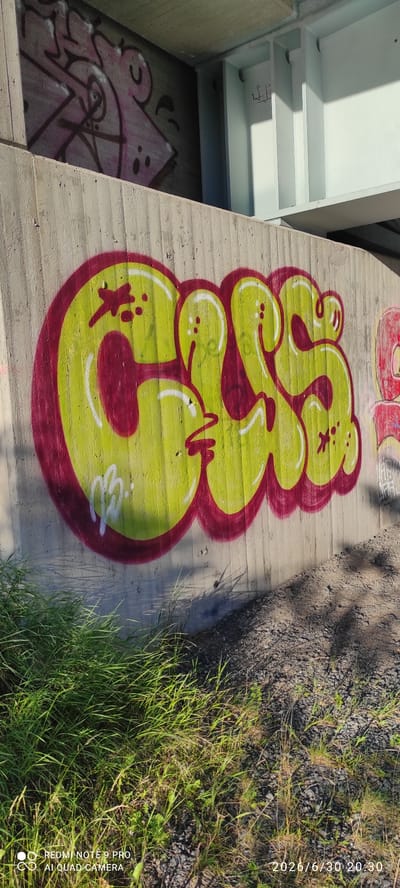 The image shows a graffiti artwork on a concrete surface. The graffiti is large, bubble-style lettering in yellow with a dark red outline and white highlights. The lettering appears to be abstract and possibly a word or tag, with flowing, rounded shapes. There are smaller details within the graffiti, such as red dots and a small red "X" shape. To the left of the graffiti, a white structure with shelves is visible, suggesting an underpass or industrial area. In the background and to the right, there is a patch of dry gravel and grass, with shadows cast by trees. The lighting suggests it is daytime, possibly late afternoon or early evening due to the long shadows. A watermark in the bottom right corner reads "REDMI NOTE 9 PRO AI QUAD CAMERA" and a timestamp "2026/6/30 20:30". The overall impression is an urban or semi-urban outdoor scene, likely a public space where graffiti is common. The graffiti itself is the main subject, rendered in a vibrant and playful style.