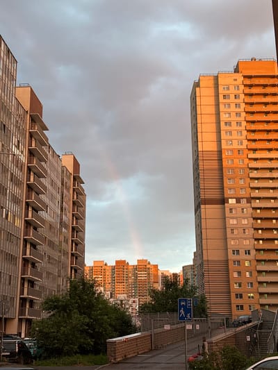 This is a vertical, eye-level, medium shot of apartment buildings in Kudrovo, Russia, under a cloudy sky. The buildings are tall and constructed with a mix of brick and concrete. The building on the left features a repetitive design with rows of balconies and windows, with some windows illuminated. The building on the right, seen from a different angle, is an orange-toned brick structure with many small windows. A faint rainbow is visible in the sky between the two buildings.

In the foreground on the right, a sidewalk with a metal fence borders a paved area. A blue sign with symbols of a car and a person walking is mounted on a pole. Beyond the fence, a person is walking, and there are several trees and some parked cars. The time of day appears to be late afternoon or early evening, as indicated by the warm light on the orange building and the soft, diffused lighting in the sky. The overall mood is somewhat somber due to the cloudy weather, but the subtle rainbow offers a touch of hope. There is some visible text on a license plate on one of the cars, but it is unreadable.