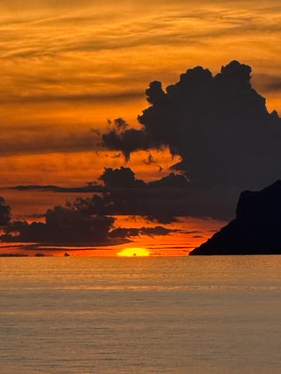 A dramatic sunset over the Gulf of Thailand illuminates the sky with vibrant orange and yellow hues. Dark, billowing clouds are silhouetted against the bright sky, with a rocky outcrop visible on the right side of the frame. The calm sea reflects the colors of the sunset, creating a serene and picturesque scene. The image likely captures a view from a beach or viewpoint on Ko Samui, Thailand, a popular tourist destination known for its stunning sunsets.