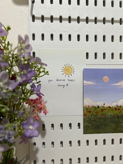 A framed photograph on a white pegboard displays a floral arrangement of purple and pink flowers in the foreground, with two cards mounted on the pegboard. The card on the left has a drawing of a smiling sun and handwritten text that reads "you deserve happy things ♡". The card on the right is a landscape painting of a field of red flowers under a light purple sky with a pale orange sun. The pegboard is mounted on a textured white wall. There is no information about the location in Semarang, Indonesia.