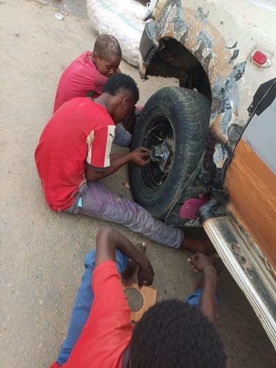 In Arawa, Nigeria, three young individuals are engaged with a damaged vehicle. Two of the individuals are crouched near the rear wheel of the vehicle, appearing to be working on it. One of these individuals wears a red t-shirt and grey pants, while the other is partially visible wearing a red shirt and blue pants. A third individual, wearing a pink t-shirt, sits further back. The vehicle itself shows signs of significant damage, particularly to the fender and wheel arch, with peeling paint and exposed metal. The ground is a rough, dusty surface, consistent with a road in a Nigerian setting. A large, white sack, possibly containing produce, is visible in the background, suggesting a market or roadside setting. The overall scene depicts a hands-on, informal interaction with a vehicle in a common urban or semi-urban environment.