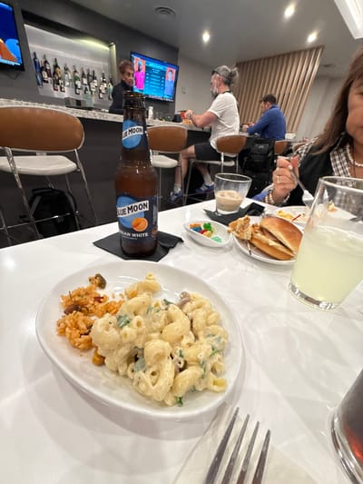 This media file captures an indoor scene, likely a bar, lounge, or restaurant, with several people dining and drinking. The primary focus is on a white table laden with various food and beverage items.
In the foreground, a white plate holds a serving of creamy macaroni and cheese with green flecks, alongside a small portion of reddish-brown rice. A bottle of "BLUE MOON BELGIAN WHITE" wheat ale, with an orange slice depicted on its label, rests on a black napkin beside the plate. A silver fork is visible on the right.
Further up the table, a partial view of a person with dark hair, wearing a plaid shirt and pearl necklace, is seen, holding a fork. Next to them is a glass containing a light yellow beverage, possibly lemonade. Another small plate holds a burger or sandwich and fried food, while a tiny white dish contains colorful gummy candies. A glass with a lighter colored drink is also visible.
In the background, several individuals are present. A man with grey hair tied in a bun and wearing a white t-shirt and grey headband is seen leaning forward. Behind him, a man in a blue shirt is visible. To the left, a bar area is visible with brown-seated stools and shelves stocked with bottles. Two flat-screen televisions are mounted; one displays "ROCK & SUMMER" text and images of people, while the other shows an abstract design. The overall setting appears modern and well-lit by artificial lights. The atmosphere suggests a casual dining or social gathering.
