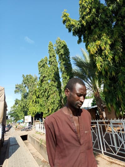 An outdoor, eye-level, medium shot shows a Black man in a brown tunic standing in front of a white metal fence. He is in the foreground, facing to his right, with his head down and eyes closed. The man has short, dark hair and a short beard. Behind him, on the right side of the frame, is a large, leafy green tree. To his left, a palm tree with a thick trunk is visible, along with more green trees behind it. In the background, the image shows a street lined with buildings and trees. A paved walkway runs along the left side of the street, and a drainage ditch is visible in the foreground. A few cars and motorcycles are parked on the street. The sky is clear and blue. This scene is located in Arawa, Nigeria.