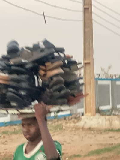 The image depicts a person in a green and white shirt. The background includes a stack of objects, possibly shoes or other items for sale, a wooden beam, a building with a white facade and gray windows, and an unpaved area suggestive of a roadside or market location. The setting is inferred to be Hayin Banki, Nigeria. The person is partially visible. The stack of items appears to be the focus. Electrical wires can also be seen.