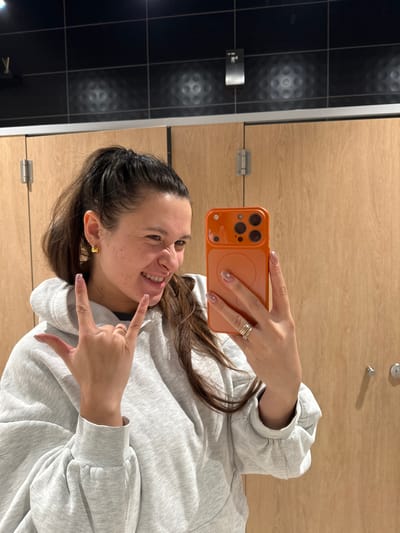 A woman takes a selfie with an orange phone in a public restroom. She is wearing a gray hoodie and has long brown hair. She makes the "rock on" hand gesture with her left hand and smiles. The background consists of black tiled walls with circular patterns and wood paneling with metal hinges and doorknobs. The photo is rotated counterclockwise.