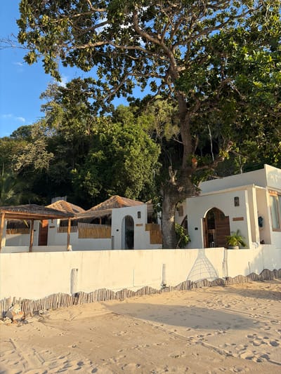 A bright, sunny day at a beach resort in Baan Tai, Thailand is depicted. The scene is dominated by a multi-story white building with arched doorways and thatched roofs, suggestive of bungalows or hotel rooms. The building is situated directly on a sandy beach, with footprints clearly visible in the sand, indicating recent human presence. A low barrier made of bamboo poles separates the resort from the beach.
Lush green trees and foliage surround the building, with a large tree with thick branches prominently featured on the left side of the image, its leaves casting shadows. The clear blue sky with a few wisps of clouds suggests pleasant weather. The time of day appears to be late afternoon or early evening, as indicated by the warm, low-angle sunlight casting long shadows.
There are no people visible in the image, but the presence of footprints and outdoor furniture on balconies suggests a resort actively being used. The overall mood is serene and tranquil, conveying a sense of a tropical getaway. No visible text is present in the image.