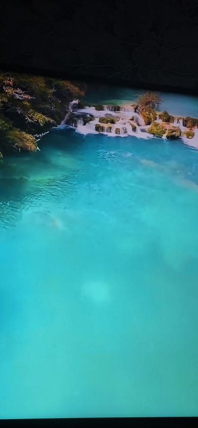The image depicts a natural scene of clear, turquoise water flowing over a series of rocky terraces, resembling a tiered waterfall. Lush green foliage and trees border the upper left portion of the scene. The water is translucent, allowing visibility of submerged rocks and ripples on the surface. The overall impression is one of a serene, natural landscape. There is no discernible indication of the city of Bila Tserkva, Ukraine, within the image. The image is a still photograph and does not contain any active elements or subjects performing actions.