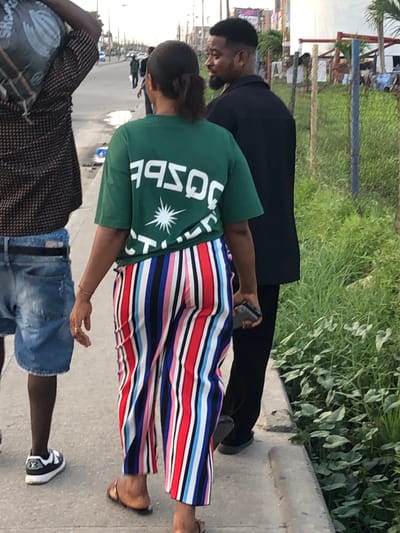 A medium shot of three individuals walking on a concrete sidewalk. One person wears a green t-shirt with white letters and a white starburst design, paired with colorful striped pants and sandals. Another person walks beside them, dressed in a black suit. The third person is wearing a brown and black patterned shirt, blue jean shorts, and black-and-white shoes. In the background, there is greenery and what appears to be a building. The setting is Alaguntan, Nigeria.