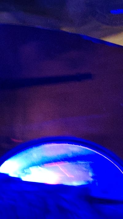 The image presents an abstract, low-light scene dominated by deep blue and purple hues. At the bottom, a prominent curved object emits a bright glow, displaying a blend of white, light blue, and distinct streaks of pink or reddish light. This illuminated element is framed by a darker blue arc, suggesting it could be a screen, a light fixture, or a reflective surface. The upper portion of the image is a textured, dark purple background, hinting at a wall or a large surface. Faint, dark, elongated shapes, possibly shadows or reflections, are discernible in the upper-middle section. The overall lighting is dim, indicating either a nighttime setting or a very dark indoor space. There are no identifiable people, objects beyond the illuminated curve, or specific activities taking place. No text is visible. The image conveys a mysterious and atmospheric mood through its play of light and shadow, with the bright, colorful illumination providing a strong focal point against the dark backdrop. The scene lacks specific location cues beyond the general context of Al-Qaboun Municipality, Syria.