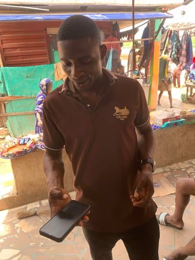 A man in a brown, ribbed polo shirt with a "CELINE PARIS" logo on the left chest, and dark trousers, stands at an outdoor market in Shagamu, Nigeria. He is holding a black smartphone in his left hand. His right hand is extended and appears to be gesturing. He is looking down at the phone. In the background, a woman in a blue and purple headscarf and matching dress is visible behind a display of packaged goods. Other stalls with clothing and various items are also present in the background. The ground is tiled with earth-toned ceramic pieces. A broom is visible leaning against a wall to the left of the man.