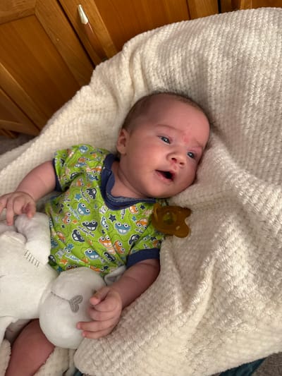A newborn baby is lying on a cream-colored blanket. The baby is wearing a green short-sleeved onesie with a pattern of cartoon cars and stars. The baby has a pacifier in its mouth. To the left of the baby is a white stuffed animal. The baby is positioned on a soft, textured surface, possibly a bed or bassinet. In the background, a wooden furniture item with a brass-colored handle is visible. This scene is not depicted within the city of Выльгорт, Russia.