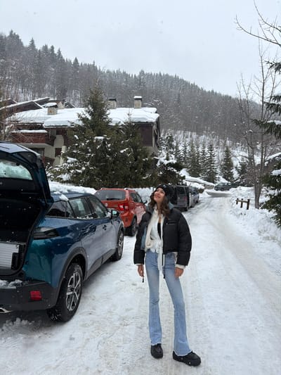 The image depicts a snow-covered scene in Les Allues, France, under an overcast sky. A young adult female stands on a snow-covered road, looking upwards with a slight smile. She wears a black beanie, a black cropped puffer jacket with white lining, a white tied shirt, light blue flared jeans, and black chunky boots. To her left, a dark teal SUV with an open rear tailgate is parked on the snowy road, revealing a silver rectangular object inside its cargo area. Behind this vehicle, an orange Suzuki Ignis and other dark vehicles are parked. In the background, multiple chalet-style buildings with snow-covered roofs and chimneys are visible, nestled amidst dense, snow-laden evergreen and deciduous trees that cover a sloping hillside. Snowflakes are discernible in the air. The ground is uniformly covered in fresh snow, with tire tracks visible on the road.