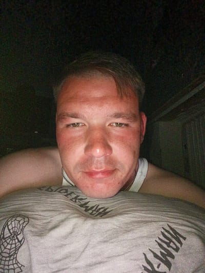 The image is a close-up, low-light selfie of a man. He is positioned in the foreground, with his face taking up a significant portion of the frame. He has fair skin, short brown hair, and light-colored eyes. He is wearing a white tank top and a silver chain necklace. His gaze is directed towards the camera. His body is partially visible, with his arms around himself or someone else, whose shoulder is visible behind his head. The background is dark and out of focus, suggesting an indoor setting at night or in low light conditions, possibly a residential area or a nondescript outdoor space with some structure visible on the right. The image does not contain any specific landmarks or features that would allow for precise localization to a specific area within Türkmenbaşy, Turkmenistan. The overall impression is that of a casual, personal photograph taken in a private setting.