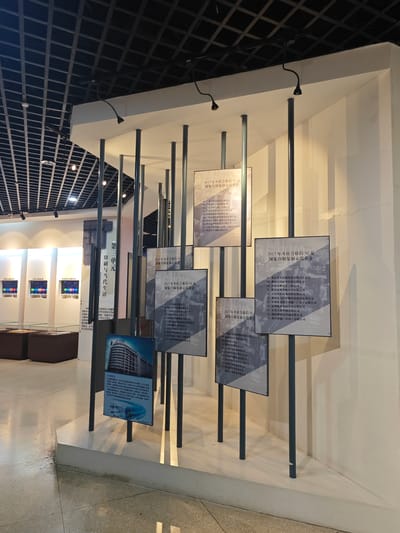 The image shows a display in an indoor setting, potentially a museum or exhibition hall, located in Daxing District, China. The display consists of several white panels with black metal poles supporting them. Each panel holds a printed sign, written in Chinese, listing organizations recognized as "National Printing and Reproduction Demonstration Enterprises" for the year 2017. The signs are illuminated by small spotlights attached to the top of the panels.

In the background, there are additional displays and seating arrangements, suggesting a larger exhibit area. The ceiling has a grid-like structure with visible lighting fixtures. The flooring appears to be polished, reflecting some of the ambient light. The overall setting is clean and well-lit.