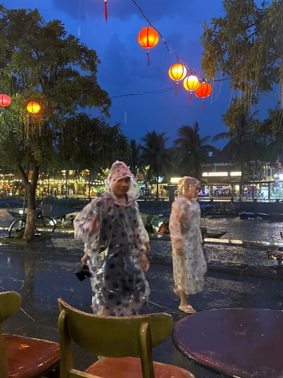 The image depicts two people wearing translucent raincoats with polka dot patterns. They are standing on a wet, paved surface, likely a street or walkway, with reflections of lights visible on the ground, suggesting it has been raining or is currently raining. In the background, there is a scene of a bustling street or market at dusk or night, illuminated by numerous bright lights from shops and stalls, with palm trees and foliage visible. Several orange, spherical lanterns are strung up on wires, also lit and adding to the festive atmosphere. The overall scene suggests an outdoor setting in Hội An Ward, Vietnam, during an evening with rain, creating a somewhat magical and reflective ambiance. The subjects appear to be interacting with the environment, perhaps observing the surroundings or walking through the rain. The presence of tables and chairs on the right side of the image indicates a seating area, possibly for an outdoor cafe or restaurant. The lighting, time of day, and the lanterns strongly suggest a vibrant, possibly tourist-oriented, location in Hội An, known for its atmospheric night markets and lantern-lined streets.