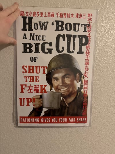 A satirical poster, likely a reproduction, is displayed in a clear plastic sleeve against a light-colored, textured wall. A human hand, with a light skin tone, is positioned in the upper left corner, partially obscuring the top edge of the poster and its sleeve. The poster's central image is a smiling Caucasian male wearing an olive-drab M1 helmet and a dark military-style shirt, holding a tarnished grey metal cup in his right hand. Large, distressed black text across the poster reads "How 'BOUT A NICE BIG CUP OF SHUT THE F𠈴K UP!", where the "U" in "SHUT" and the "CK" in "FUCK" are replaced by Japanese characters. Smaller red text at the bottom states "RATIONING GIVES YOU YOUR FAIR SHARE". Additional red Japanese characters are arranged vertically along the right side and horizontally at the top of the poster. The wall exhibits a consistent, rough stucco-like texture. This scene is located in the Town of Vestal, United States.