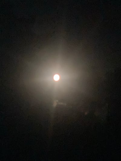 A full moon is visible in the night sky, surrounded by a hazy glow. The moon is centrally located in the frame, appearing as a bright, circular orb against the dark, textured background. The lower portion of the image shows faint, indistinct shapes that may be trees or buildings silhouetted against the moonlight. There is a noticeable amount of digital noise or grain throughout the image. The scene appears to be an outdoor night shot, likely taken from Batak, Bulgaria, given the prompt. No people or specific actions are depicted.