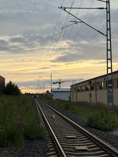 The image depicts a railway track in Pori, Finland, during what appears to be either sunrise or sunset, with a cloudy sky tinged with yellow and orange hues. The track runs from the foreground into the distance, flanked by lush green vegetation on the left and industrial buildings on the right. Overhead, a complex network of catenary wires for an electric railway is visible, supported by metal poles.
To the left of the tracks, there is dense green foliage with some small pink flowers. In the distance, a large industrial building with a light-colored facade and windows is prominent. Adjacent to it, a construction crane is visible, suggesting ongoing development or activity in the area. A dark-colored building is also present on the far left.
On the right side of the image, a more detailed view of the industrial building reveals yellow siding and windows. Several signs are attached to a metal pole next to the building, including a yellow triangular sign with a black arrow pointing downwards and a yellow rectangular sign with a black "E" inside. Another sign, labeled "324-6" and "276", is also affixed to the pole. The ground surrounding the tracks is covered with dark gravel. The overall scene is one of an industrial area bordering a railway line under a dramatic sky.