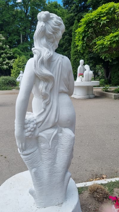 The image displays a white statue of a woman from behind, seen in a park setting. The statue depicts a woman with her hair styled in an updo and long, flowing curls. She is draped in fabric, and her left arm is bent, holding a bunch of grapes. The statue appears to be made of concrete and is mounted on a white pedestal. In the background, there are trees, other statues, and a paved walkway. Further in the background, another white sculpture of two figures leaning towards each other can be seen. The overall scene is of an outdoor public space, likely a garden or park. The context of Smila, Ukraine is not visually apparent from the image itself.