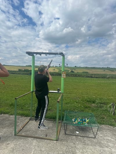 The image captures a person aiming a shotgun at a clay pigeon shooting range. The subject is a young person wearing a black t-shirt, black pants with white stripes, and a green baseball cap. They are positioned within a metal frame structure, holding the shotgun to their shoulder. In the background, a green field stretches towards a distant tree line and agricultural land. The sky is partly cloudy with a mix of blue patches and large, grey clouds.
To the right of the frame, a green metal cage contains numerous colorful clay pigeon targets, mostly blue, yellow, and white. A small, brown sign with letters and symbols is attached to the green metal pole in the foreground. A concrete surface surrounds the shooting stand. The overall scene suggests an outdoor recreational activity under a cloudy daytime sky. The location is identified as Фельдмаршальский, Russia.