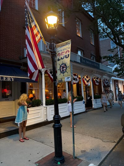 The image captures a street scene in the Village of Sag Harbor, United States of America, during twilight. A brick building, identified as "The American Hotel" by signage, dominates the background, adorned with red, white, and blue star-spangled bunting. An American flag is prominently displayed on a flagpole to the left of the hotel entrance. In front of the hotel, a sidewalk runs along the street, with several people walking.

To the left of the hotel, an entrance labeled "GALLERY" is visible, with a blue awning overhead. A young woman with blonde hair, wearing a light blue dress and red flip-flops, stands on the sidewalk facing away from the camera, looking towards the hotel entrance. Further down the sidewalk, a group of people are walking, including a woman in a short light-colored dress, a man in a light-colored shirt and shorts, another man in a light-colored shirt and shorts, a woman in a dark top and shorts, and a woman in a dark patterned top and shorts.

A lamppost with an illuminated lamp stands in the foreground. A banner hanging from the lamppost displays "CELEBRATING AMERICA 250" along with a nautical symbol and text related to Sag Harbor. The street is paved, and the sidewalk is made of concrete. Trees with dark green foliage are visible, suggesting an outdoor setting. The lighting indicates it is either dusk or early evening.