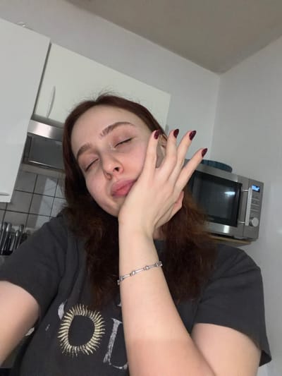 A woman with brown hair and closed eyes covers part of her face with her hand, revealing burgundy nails. She wears a silver bracelet. She appears to be in a kitchen with a white refrigerator to the left and a stainless steel oven in the background. Her dark grey t-shirt features a circular graphic. The picture has a slightly rotated perspective.