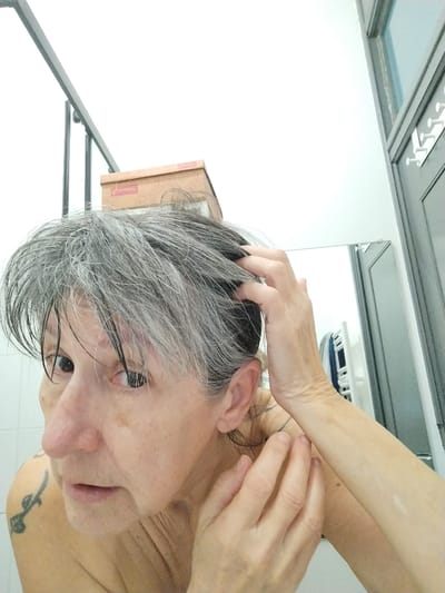 The image captures a female individual, appearing middle-aged to elderly, with wet, disheveled grey hair, looking directly towards the viewer. Her left hand is positioned within her hair, suggesting an action of ruffling or drying. Fair skin is observed on her face, displaying multiple brown macules. A dark-inked tattoo of an unspecified design is present on her upper left shoulder. The subject is bare-shouldered. The background indicates an indoor environment, consistent with a bathroom, featuring white tiled walls on the left. To the right, a mirror reflects a white, bar-style towel rack holding blue and white fabric items. A dark-framed structure, possibly a doorway or shower enclosure, equipped with white wall hooks, is visible on the far right. Above the subject's head, a dark horizontal metal bar is present. A brown cardboard box with the label "CHARDON" is partially visible in the upper left background. The image contains no explicit visual indicators to verify the location as Montélimar, France.