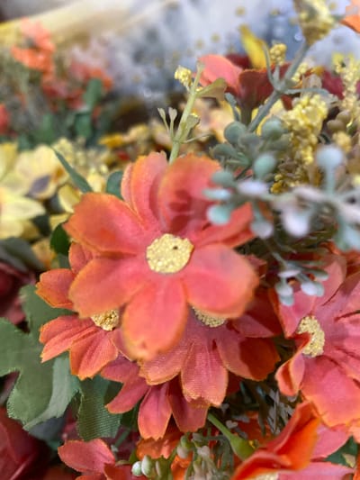 A close-up, slightly blurred, overhead shot displays a bouquet of artificial flowers in warm tones of orange, yellow, and red. The central focus is on several orange daisy-like flowers, each with delicate, textured petals and yellow centers. Green leaves and stems are interspersed throughout the arrangement, along with clusters of tiny yellow flowers and some fuzzy, pale green buds. The background is out of focus, suggesting a shallow depth of field, with hints of lighter colors that could be other flowers or a decorative element. The lighting is soft, creating a gentle illumination of the floral subjects. The scene is devoid of people and action, presenting a still-life composition. This image does not depict Giginyu, Nigeria, or any specific urban environment.