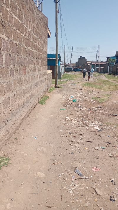 The scene depicts an unpaved dirt road in Mosop ward, Kenya, under a clear blue sky with bright daylight. A textured stone block wall runs along the left side of the frame, bordered by a narrow dirt path and sparse green grass. A wooden utility pole, supporting multiple overhead wires, stands centrally alongside the wall. The dirt road itself is extensively littered with various discarded items, including plastic bags, paper, and miscellaneous debris. In the mid-ground, three adult individuals are observed walking away from the camera down the road. Further into the background, a series of low-rise buildings constructed from masonry and corrugated iron are visible, interspersed with additional utility poles. A blue pickup truck and a dark SUV are parked on the right side of the road in the distance. No direct interactions between the walking individuals are discernible.