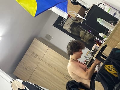 A shirtless young man with curly brown hair is sitting at a desk in a room. He is wearing black shorts and a beaded necklace. The man is looking down and appears to be interacting with something on the desk. The desk is cluttered with various objects, including a computer monitor, bottles, and what appears to be vape juice. Behind the desk, a black banner with white text and imagery of a forest and a figure is hanging on the wall. Above the banner, a Ukrainian flag is draped. In the background, a light-colored wooden wardrobe with silver handles is visible. The room appears to be a bedroom or study. The lighting suggests it could be daytime. There is no indication of the weather.