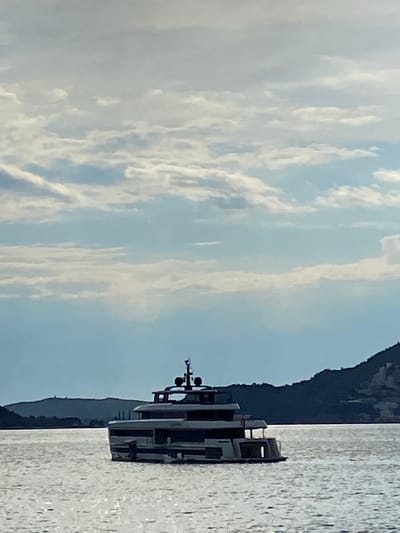 A horizontal image depicts a yacht on water, likely the Adriatic Sea, with a mountainous coastline on the right side and a partly cloudy sky to the left. The yacht is gray and boxy in shape. The water is reflective. The location is probably Pržno, Montenegro based on the geography and coastal features, specifically the mountain shoreline in the background and the presence of yachts in the area. No people are visible.