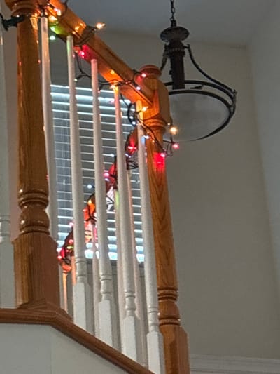 A portion of a staircase is decorated with colorful string lights. The railing, made of light brown wood, features white balusters. A black light fixture with a circular shade hangs overhead. The background wall is a neutral light gray. Light streams through the blinds behind the railing, illuminating the stairs.