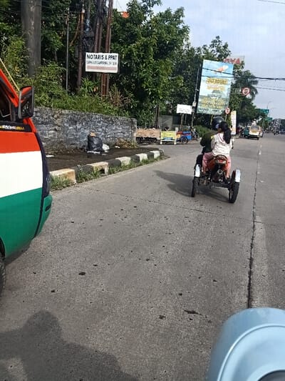 A motorized three-wheeled vehicle travels down a grey asphalt road. It carries two people, with the rear passenger wearing a patterned jacket and a helmet. The vehicle casts a shadow on the road. To the left, a colorful van is partially visible. In the background, lush green trees line the road, punctuated by signs, including one for "NOTARIS & PPAT." Other vehicles are further down the road. A blue object appears at the bottom right corner.