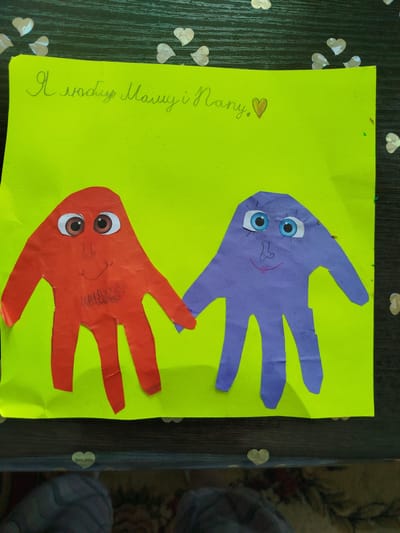 The image shows a craft project on a bright green piece of paper, laid on a dark wooden surface decorated with small white heart-shaped confetti. The craft features two handprints cut out of construction paper and pasted onto the green paper. One handprint is red, and the other is purple. Both handprints have been transformed into characters with googly eyes, drawn noses, and smiling mouths. The red hand has a drawn beard, while the purple hand has drawn eyelashes. Above the handprints, in handwritten cursive, is text in Ukrainian. The red hand is on the left, and the purple hand is on the right, with their fingers intertwined. The scene appears to be indoors, likely a home or classroom setting.