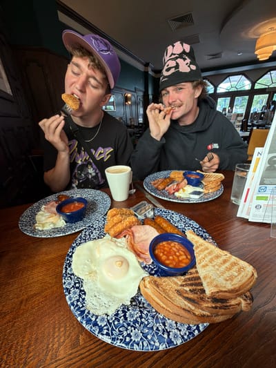 Two men are seated at a wooden table, presumably dining. One man is wearing a black hooded sweatshirt and a beanie, while the other wears a black T-shirt and a purple baseball cap. They are both holding food on forks and smiling. On the table are three plates of food, each containing a Full English breakfast including toast, baked beans, bacon, eggs, sausage, and hash browns. Also on the table is a white mug or cup. Above them are ceiling fixtures, and in the background, a window and interior of what appears to be a restaurant or pub. There is a stack of papers or newspapers with the letter "Z" visible on the top. The location is Reigate and Banstead, United Kingdom.