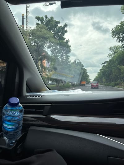 The image captures a view from inside a car, looking out the front windshield on an overcast day in Surabaya, Indonesia. The sky, filled with soft, diffused clouds, dominates the upper part of the scene. Lush green trees line both sides of the road ahead, creating a canopy effect.
On the road, a red car is visible in the distance, followed by what appears to be a yellow or orange vehicle, suggesting traffic. To the lower left, a decorative metal structure, possibly a street light or signpost, can be seen partially.
Inside the vehicle, the dark-toned dashboard and air vents are prominent in the lower and right sections of the frame. A label reading "AIRBAG" is visible on the passenger side of the dashboard. On the windshield, a small sticker with "3M" is discernable. A plastic water bottle, with "AQUA" (reversed) and "1624" printed on its label, rests on the dashboard near the center console. The car interior appears modern and well-maintained. No people are visible in the image. The scene suggests travel during daylight hours, under cloudy weather conditions.