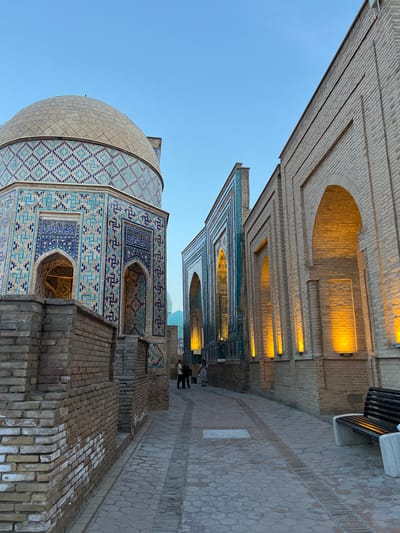 The image depicts a narrow walkway between two intricately decorated buildings, likely a religious or historical site in Samarkand, Uzbekistan. The architecture features prominent use of light-colored brickwork, with arched openings and decorative friezes. One building, on the left, displays a large dome and walls adorned with vibrant blue and white geometric tilework. The other building, on the right, showcases similar tilework around its arched windows, with warm light emanating from within, suggesting interior illumination. The walkway is paved with stone and brick, creating a textured surface. In the distance, a few people are visible walking along the path, appearing small in the grand scale of the architecture. A bench is situated against the wall of the right building, providing a resting place. The clear blue sky above indicates it is daytime, likely late afternoon or early evening, given the warm glow of the interior lights. The overall atmosphere is one of historical grandeur and architectural beauty. No visible text is present in the image.