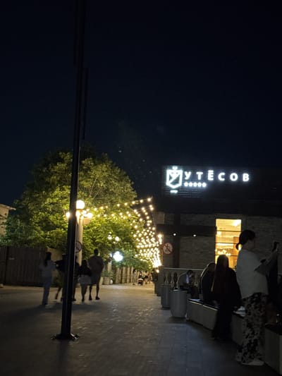 The image depicts a nighttime scene in Anapa, Russia, with a pedestrian walkway illuminated by string lights draped over trees and a street lamp. A building with a prominent sign reading "УТЕСОВ" is visible in the background, featuring stylized letters and a crest-like emblem. Several individuals are walking and sitting along the pathway. On the left, a group of people are strolling, with one person in a white top and light-colored pants in the foreground. To the right, a woman in a white top and patterned pants stands, appearing to hold a phone. Other individuals are seated on benches. The ground is paved with cobblestones, and concrete planters line the walkway. A lamppost stands in the foreground, partially obscuring the view.
