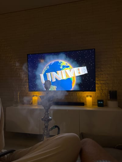 The image captures a dimly lit room with a large television screen displaying the Universal Pictures logo. The logo features a globe surrounded by stars, with the word "UNIVERSAL" emblazoned across it in large, white, three-dimensional letters. The television is mounted on a wall covered in textured, light-colored bricks. To the right of the television, a white cabinet or dresser is visible, with a dark digital clock displaying "22:25" and two illuminated yellow lamps. In the foreground, a hookah with smoke billowing from it is positioned in front of the television, partially obscuring the lower part of the screen and the cabinet. The presence of the hookah suggests a relaxed or social gathering. The overall ambiance is cozy and somewhat dimly lit, suggesting it is nighttime. There are no people clearly visible, though a person's legs and arm are partially in the frame on the far right, draped in light-colored fabric. The scene appears to be indoors, in a residential setting. The visible text is "22:25" on the clock and "UNIVERSAL" on the television.