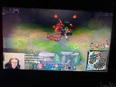 The image captures a monitor screen displaying a video game, positioned within an indoor environment in the city of Yambol, Bulgaria. The immediate surroundings visible are a dimly lit room, with minimal additional detail discernible beyond the monitor itself. The monitor prominently features the user interface and gameplay of League of Legends. In the upper left corner of the game screen, a health bar indicates a character with 1185/1256 health and 457/513 mana. Combat is depicted in the central area, showing multiple characters engaged in an interaction, with health bars indicating damage. Red-colored minions are visible advancing on the left side of the screen, near larger character models. Floating red-orange visual effects are present in the upper-mid section of the screen, suggestive of in-game spell effects or items. A chat log is displayed on the bottom left, featuring time-stamped messages such as "06:11 doki doki hoes (Shaco) is asking for assistance," "06:15 stability (Janna) is on the way," "06:17 stability (Janna): Pantheon - Alive," and "06:25 permalimittest (Lucian) is on a killing spree!". Below the game screen, in the bottom left corner, a live camera feed of a male individual is visible. He is wearing glasses and a black hoodie, with a headset positioned over his ears. He is seated in a gaming chair, looking directly forward. To the right of his feed, a game overlay shows chat messages from users like "vicstasia," "Spaghetti15," "ezattack420," and "sxorn1." Below this, player statistics are displayed, including a score of 124 minion kills, 45 ability power, 0.79 attack speed, and 0% critical strike chance. The player's gold count is 481. The lower central portion of the screen displays the player's in-game character abilities, showing cooldowns and mana costs, alongside items purchased. A health bar indicates 1185/1435 health and 571/776 mana, with regeneration rates of +10.9 health and +2.2 mana. On the right side of the game screen, a minimap of the game's terrain is displayed, showing character positions, objectives, and areas under vision control. Above the minimap, small portraits of allied champions are visible, indicating their current health and status. The upper right corner of the game screen displays "FPS: 229" and "21 ms," indicating performance metrics. A white cursor is visible near the top right of the minimap, and another white cursor is visible mid-screen, near the center of the gameplay action. The overall scene is well-lit from the screen's output, contrasted