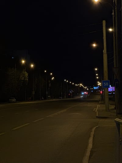 The image depicts a nighttime scene on a street in Tyumen, Russia. The street is illuminated by numerous streetlights, casting a warm glow on the asphalt. Several cars are present on the road, including a silver sedan parked on the left side. A few people are visible walking on the sidewalk on the left side of the street, and a motorcycle is parked further down. Traffic lights are visible in the distance, and a speed limit sign indicating "50" is prominently displayed on a pole on the right side of the street, along with a directional sign. Trees line the left side of the road, and a building is partially visible in the background. The overall atmosphere is quiet and dimly lit, characteristic of a city street at night.