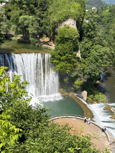 This image captures a breathtaking view of the Pliva Waterfalls in Jajce, Bosnia and Herzegovina. The scene is dominated by lush greenery and cascading water, suggesting a natural and serene environment. The waterfall is the primary subject, a wide curtain of white water tumbling down a rocky formation into a pool of calm, turquoise water below. The sunlight catches the spray, creating a luminous effect.
On a curved concrete embankment adjacent to the pool, three individuals are visible. One person, wearing a blue top, is seated, while two others, in white tops, appear to be standing or leaning. They are positioned on the embankment, observing the waterfall and the surrounding landscape, perhaps enjoying the view or taking a break. The embankment itself is part of a man-made structure that seems designed to enhance the viewing experience of the natural wonder.
Further in the background, more of the waterfall's cascades are visible, interspersed with rocky ledges and dense foliage. The overall impression is one of natural beauty combined with thoughtful integration of human elements for appreciation. The time of day appears to be sunny, with the bright light suggesting midday or early afternoon. The lush trees indicate a warm season, likely spring or summer. The scene evokes a sense of peace and wonder, with the power and beauty of nature on full display. No visible text is present in the image.