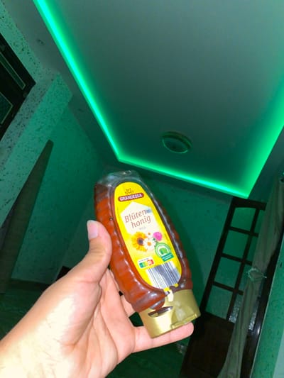 A person's hand holds a bottle of Grandessa Blutenhonig (flower honey) in a room with green ambient lighting. The honey bottle is plastic with a gold cap and a yellow label featuring images of sunflowers and other flowers. The background shows a textured green wall and a ceiling with a green LED strip light. A dark-framed window or door is visible to the right. The overall lighting is dim, with a strong green hue. The scene appears to be indoors, possibly in a residential or commercial space in Baraki, Algeria, given the context provided.