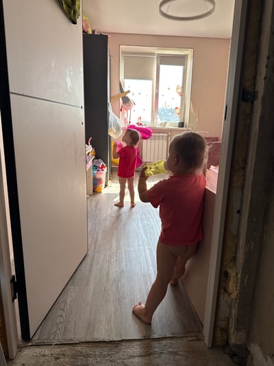 A vertically oriented photograph taken indoors shows two young children in a room that appears to be undergoing renovation. In the foreground, a white cabinet dominates the bottom half of the frame. Behind it, a dark cabinet is visible, stocked with various toys in a clear plastic bin.
To the right of the dark cabinet, a young child, dressed in a pink t-shirt and shorts, is standing and holding a yellow cup to their mouth. Further to the left and slightly behind this child, another child, also in pink, is lying on the floor.
The room has light pink walls and a window with a view of an urban setting, suggesting it's daytime. The floor is covered in wood-look laminate. The ceiling area above the doorway shows exposed insulation and rough concrete, indicating unfinished construction. The overall scene suggests a domestic setting in Chelyabinsk, Russia, where everyday life continues amidst renovation work.