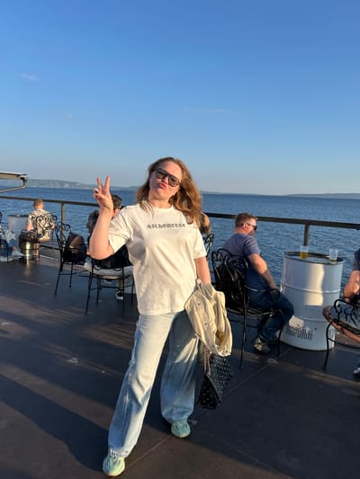 A young woman with blonde hair and sunglasses poses for a photo on a boat, holding up two fingers in a peace sign. She is wearing a white t-shirt, light-wash jeans, and light blue sneakers. Next to her is a black studded purse and a light-colored jacket. In the background, two men are seated on a boat deck, looking out at the water. One man, wearing a blue t-shirt and jeans, is reclining in his chair. Another man, in a patterned shirt, is seated further back. A white barrel with two glasses of amber liquid on it serves as a table. The scene is set on a sunny day, with a clear blue sky and calm water. The boat appears to be on a body of water with hills or land visible in the distance. Text on the white barrel is partially visible and appears to be in Cyrillic, possibly related to a brand or location.