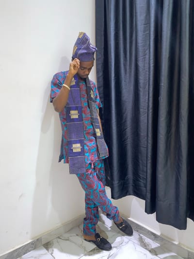 A man in vibrant blue and red patterned Yoruba attire is seen reclining. He wears a matching cap and suede loafers. A sash with blue and metallic patterns adorns his upper body. A dark, draped curtain forms a backdrop. The setting is indoors, suggested by a white wall and marble-tiled corner.